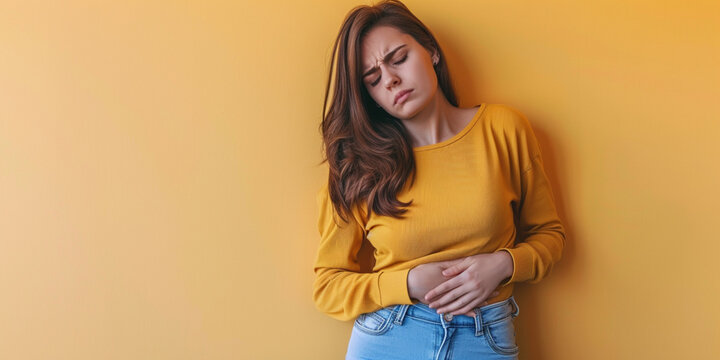 Indoor shot of displeased woman suffering from painful period cramps, holds hands on belly, has pain in stomach, stomachache, upset expression, wears long sleeve shirt on yellow background