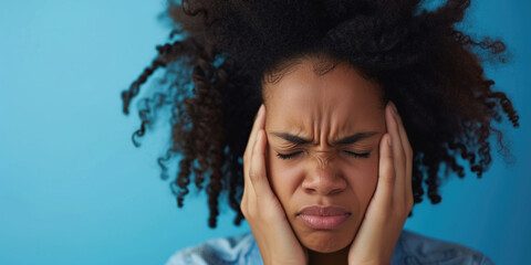 African American young woman holding hands near head while suffering from painful headache migraine on studio blue background. Woman suffer from stress or a headache grimacing in pain - obrazy, fototapety, plakaty