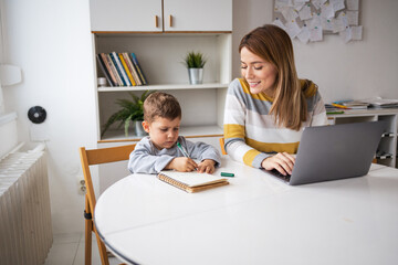 Happy mother using laptop sitting beside son doing homework at desk