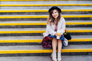 Portrait of young laughing fashion woman with fresh tulips in net bag holding reusable coffee cup while sitting on stairs at the city street. Urban street fashion. Springtime mood. Selective focus.