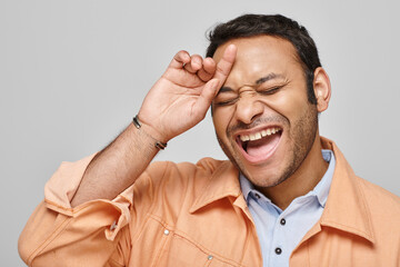 cheerful handsome indian man in orange vibrant jacket laughing hard and posing on gray background