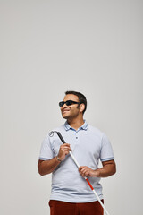 joyous indian blind man in blue tee shirt with glasses and walking stick posing on gray backdrop