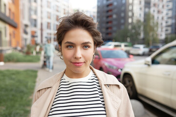 Happy young female real estate agent in stylish trench standing against multi-storey buildings and parked cars in neighborhood or city residential area, smiling at camera, on her way to make deal