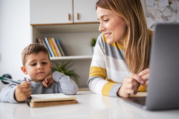 Working mother with laptop looking at son doing homework at home