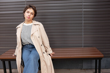 Outdoor image of stylish trendy confident girl with short haircut in beige trench and jeans sitting on bench against brown wall with copy space. Female fashion, spring outfit concept