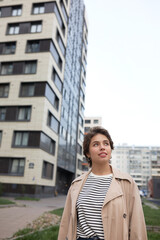 Outdoor image of attractive stylish female real estate agent standing in new residential area, looking around. Pretty girl in trench searching for good apartment to buy in comfortable neighborhood