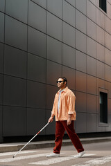 attractive indian blind man in orange jacket with glasses and stick with gray wall on backdrop