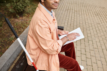 cropped view of indian man with blindness in orange jacket sitting on bench and reading braille code