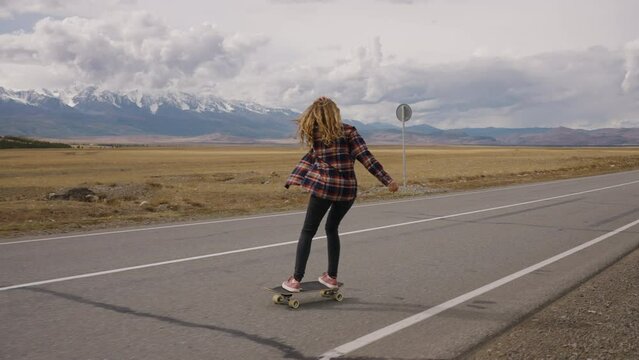 Carefree extreme girl rides a longboard in an unbuttoned plaid shirt, enjoying life. Back view of a woman skateboarding on an empty road with breathtaking mountain views under a cloudy sky