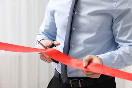 Man cutting red ribbon with scissors indoors, closeup