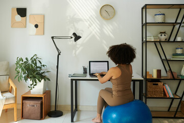 Woman sitting on fitness ball when working on laptop at home, view from behind