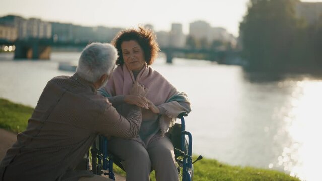 Husband walking with wife in wheelchair. Senior family couple spending time in city park near pond. Man hugging female showing love care, they smiling. Living with physical disability, caring concept.