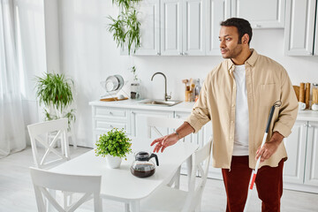 attractive indian man with blindness using walking stick and holding coffee pot while in kitchen