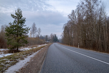 Clean winter road in forest with turns and curve with trees. Winter road with white dividing line at winter season. Melting dirty snow on road side.