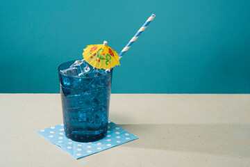Cocktail in blue glass  filled with ice with small yellow umbrella and straw on blue napkin on a white table