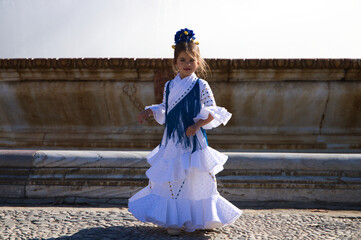 A pretty little girl dancing flamenco dressed in a white dress with ruffles and blue fringes in a...