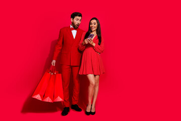 Full body photo of two elegant beautiful impressed people hold shop bags use smart phone empty space isolated on red color background