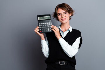 Portrait of intelligent positive girl with bob hairdo dressed waistcoat in eyewear showing calculator isolated on gray color background