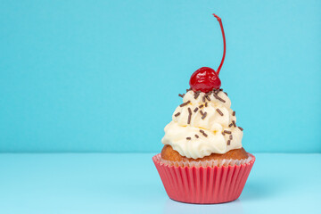 cupcake with whipped cream decorated chocolate sprinkles and maraschino cherry on blue background