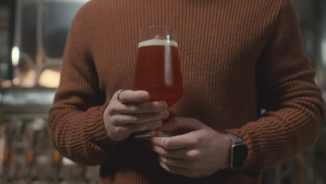 Cropped slowmo of unrecognizable micro-brewery owner wearing knitted red brick color sweater holding glass of fresh bear standing indoors with stainless steel beer production equipment in background