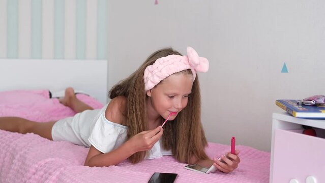 The girl lies on the bed and paints her lips. The child looks in the mirror and applies lip balm.