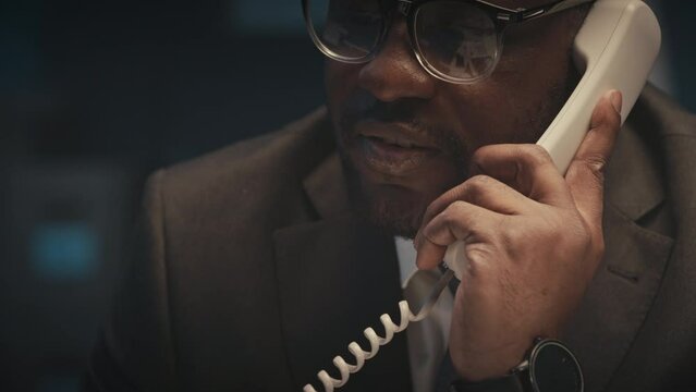 Tilt up of workaholic Black businessman dialing phone number on landline and staring phone conversation while overworking alone in office at night