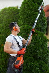 Caucasian handyman in overalls, protective glasses, and gloves pruning hedge on backyard....