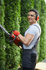 Professional caucasian gardener in uniform, safety glasses, headphones and gloves cutting hedge on back yard. Handsome man using electric trimming machine for work at garden.