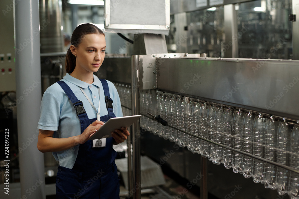 Poster young female inspector with tablet standing in front of factory conveyor and controlling quality of 