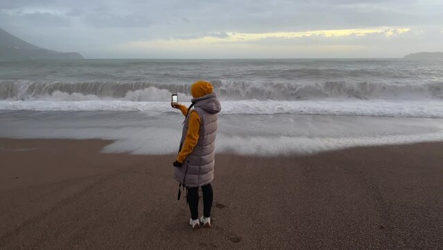 A girl standing on a sandy beach films big sea waves on her phone.