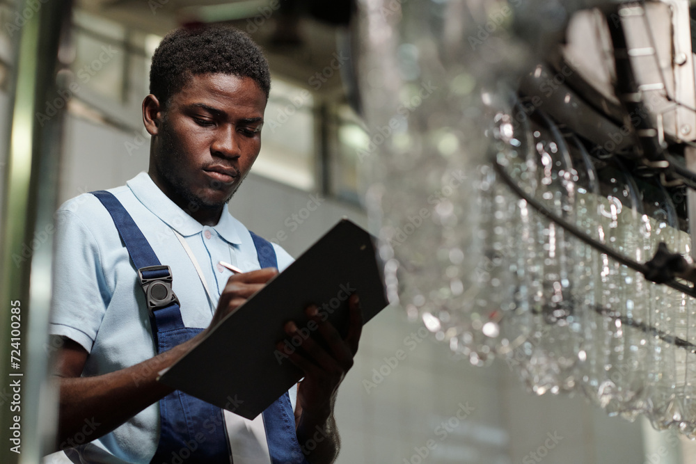 Aufkleber young male engineer of modern bottled water factory making notes in delivery paper while standing ag