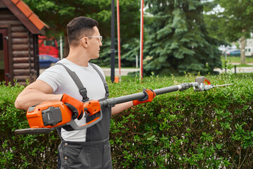 Experienced male gardener in working clothes, safety glasses and gloves using hand electric trimmer for cutting overgrown bushes. Manual work at summer garden.