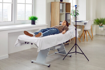 Teenage child girl lying on the couch in the clinic while receiving IV drip infusion and vitamin...