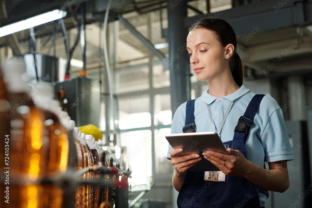 Poster Young female inspector with tablet looking at assembly line with capped bottles with drinks and checking quality of plastic packaging