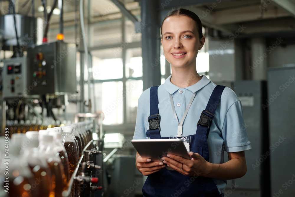 Aufkleber Young smiling female inspector with tablet looking at camera while standing by assembly line with row of capped bottles