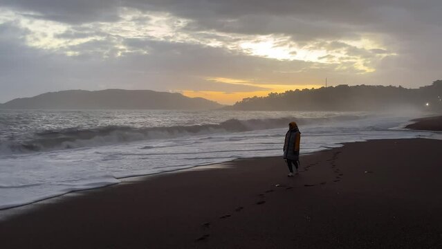 A girl walks along a sandy beach along a stormy sea against the backdrop of a beautiful sunset.