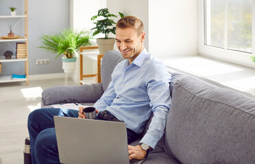 Happy young business man sitting on the sofa at home, looking at the screen of his laptop computer, watching a funny video, smiling, holding a cup from a thermos bottle and drinking hot tea or coffee