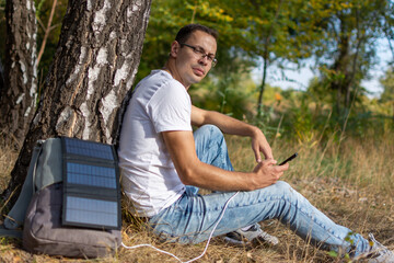 A guy in glasses sits by a tree and charges his phone with a solar panel