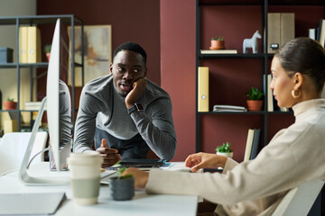 Fototapeta premium Young African American businessman bending over workplace of female colleague and looking at her during chat in coworking space
