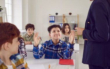 Young high school student boy raising hand to answer on a lesson sitting at the desk to a female teacher in the classroom with classmates. Education, knowledge and junior high school concept.