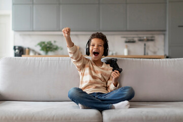 Black boy in headphones holding game controller and raising fist in victory