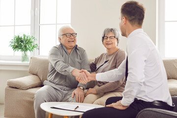 Happy smiling elderly person shaking hands with man advisor for health insurance sitting on sofa at home in retirement. Senior couple talking and make a deal with male financial agent.
