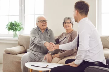 Deurstickers Happy smiling elderly person shaking hands with man advisor for health insurance sitting on sofa at home in retirement. Senior couple talking and make a deal with male financial agent. © Studio Romantic