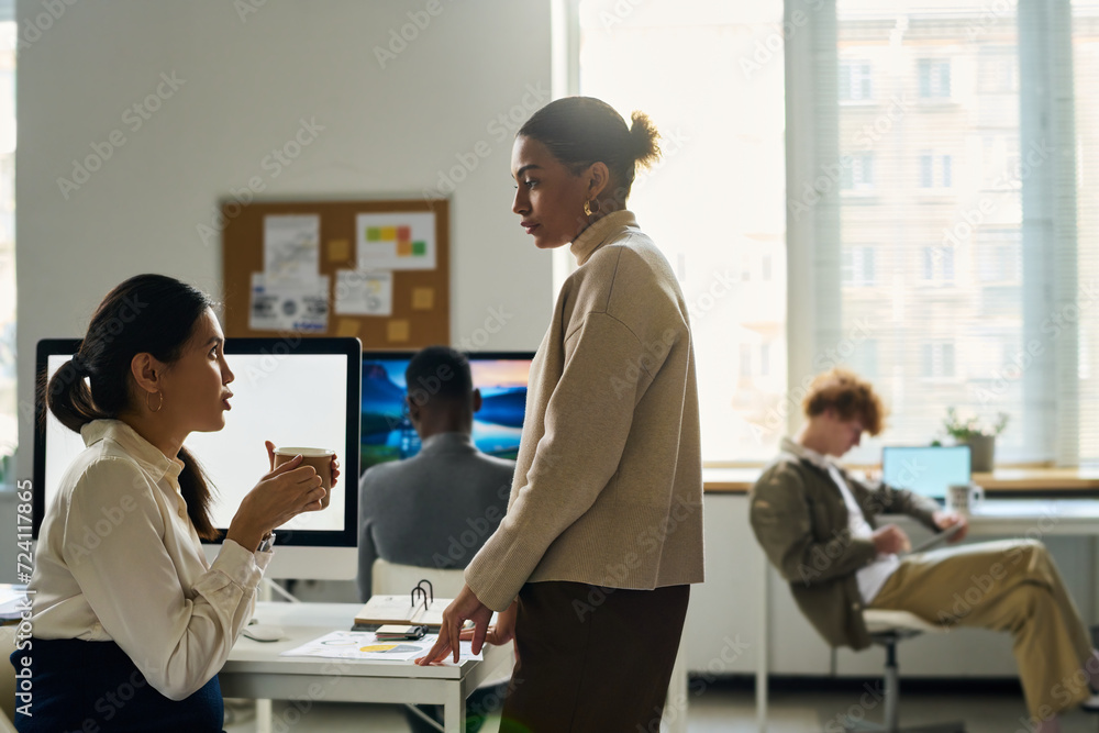 Poster side view of two young intercultural businesswomen having chat at coffee break while one of them sit