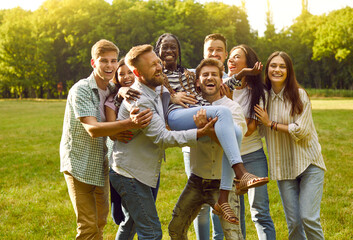 Group of friends having fun outdoors. Multiracial group of cheerful best friends having fun together enjoying summer day in park. Men and women lift up their dark-skinned girlfriend. Lifestyle concept - obrazy, fototapety, plakaty