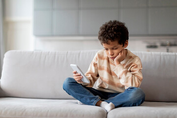 Little black boy sitting on sofa, looking disinterestedly at his smartphone - obrazy, fototapety, plakaty