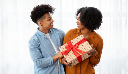 A joyful young couple shares a special moment, exchanging a beautifully wrapped gift with a red...