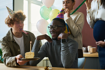 Happy young man in birthday cap looking at piece of tasty cake with burning candle while sitting by...