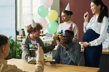 Group of happy young intercultural colleagues in birthday caps surrounding African American man...