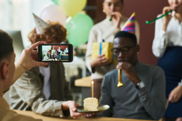 Hand of young man holding smartphone in front of group of happy colleagues celebrating birthday of...